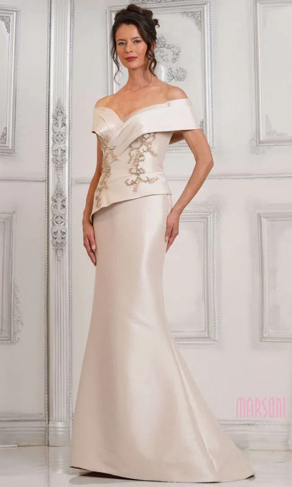 A woman wears the Marsoni MV1312 long formal dress in champagne, featuring an off-shoulder fitted stretch Mikado design with embroidered details and a mermaid skirt, posing before an ornate, light-colored wall.